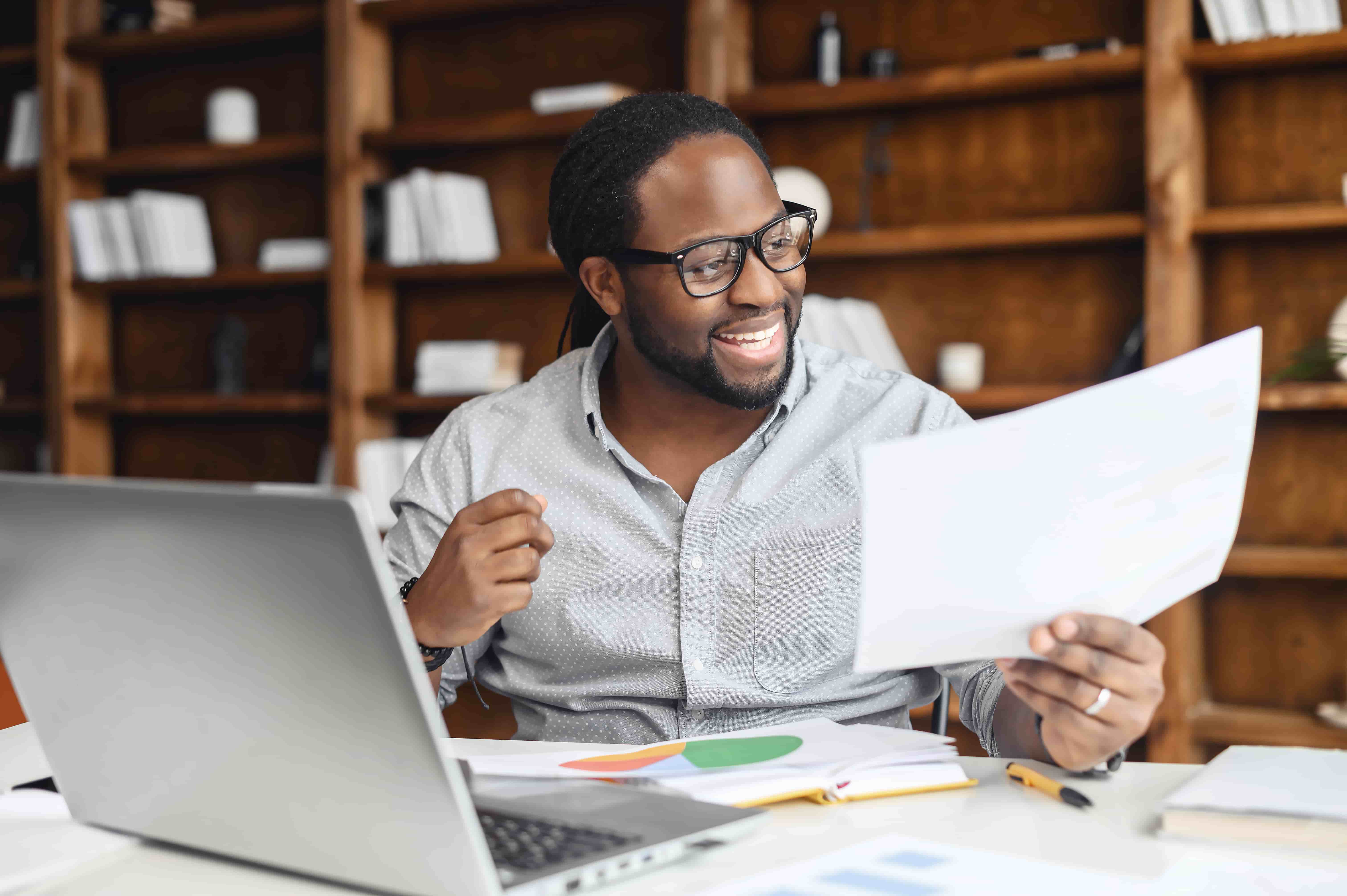 Happy African-American businessman in eyeglasses looks at a document, satisfied with report results, analyzing raising of sales graphs, doing paperwork, glad to see good project result, a laptop near
